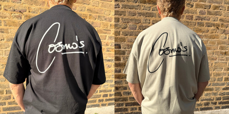 the backs of a black and a stone cosmo’s tee, each printed with the cosmo’s signature